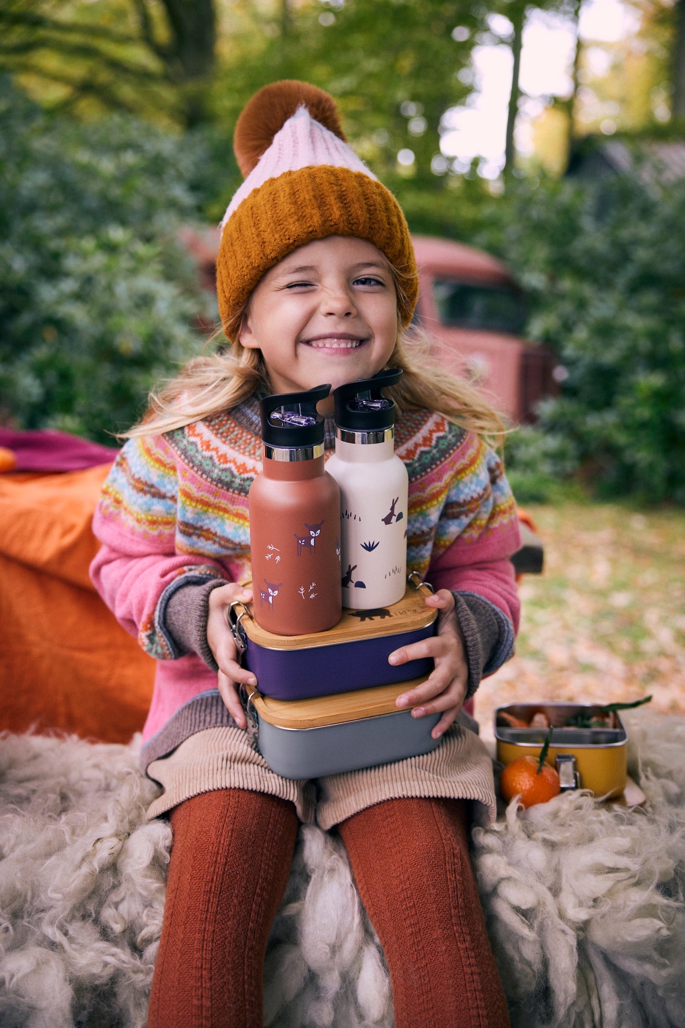 Mädchen mit bunter Kleidung beim Picknick mit Trinkflaschen und Brotdosen auf dem Schoß 